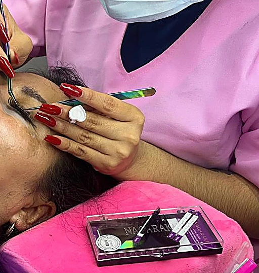Manos de la técnica con uñas rojas aplicando extensiones con pinzas, junto a una charola de pestañas sobre cojín rosa