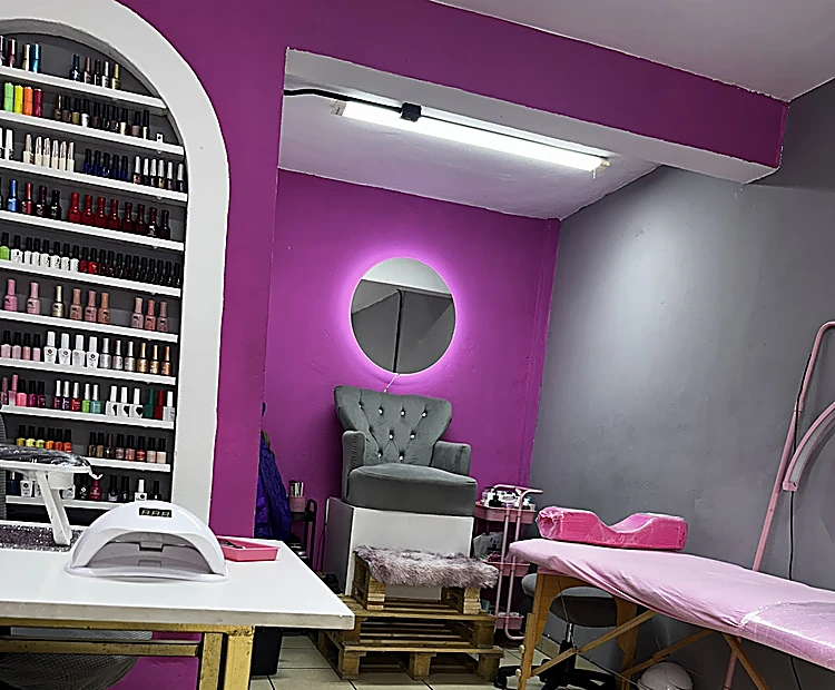 Interior: arco blanco con repisas de esmaltes por color, mesa de manicura con lámpara, muro magenta con espejo redondo iluminado y camilla rosa