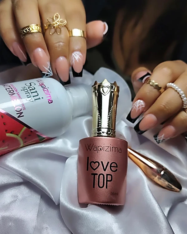 Uñas largas en francés negro con punta plateada, anillos dorados y un frasco rosa de top coat sobre satín blanco